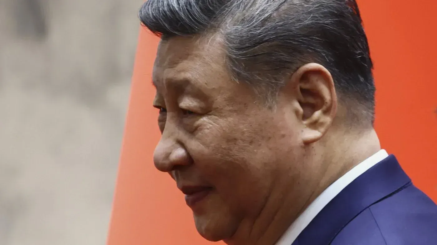 صورة جانبية لشي جين بينغ، الرئيس الصيني، يظهر فيها تعبيره الجاد أمام خلفية حمراء، مما يعكس التركيز على القضايا الأمنية والسياسية.