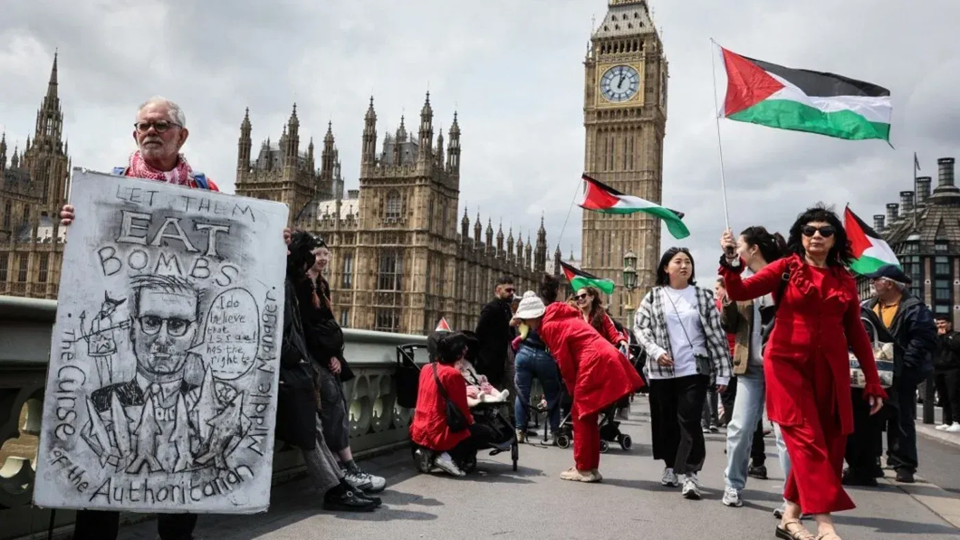 احتجاج في لندن يحمل المشاركون فيه أعلام فلسطين، مع لافتة تعبر عن موقف سياسي، وخلفهم مبنى البرلمان البريطاني.