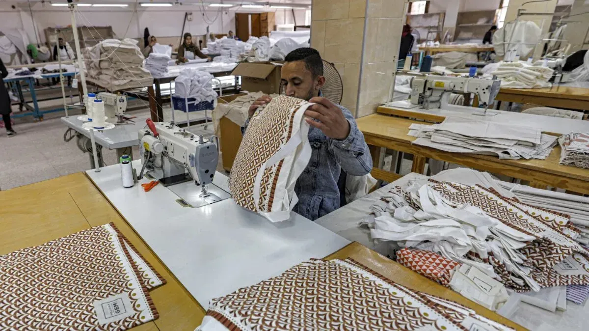 عامل مصري يقوم بخياطة الملابس في مصنع، مع وجود مواد خام وأقمشة حوله، في سياق تحديات الصناعة التحويلية في مصر.