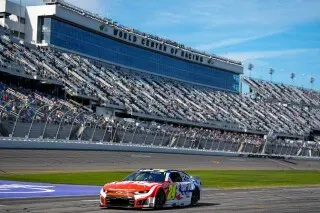 سيارة سباق شيفروليه رقم 24 لويليام بايرون تتسابق على حلبة دايتونا 500، مع مدرجات ممتلئة في الخلفية تحت سماء زرقاء.