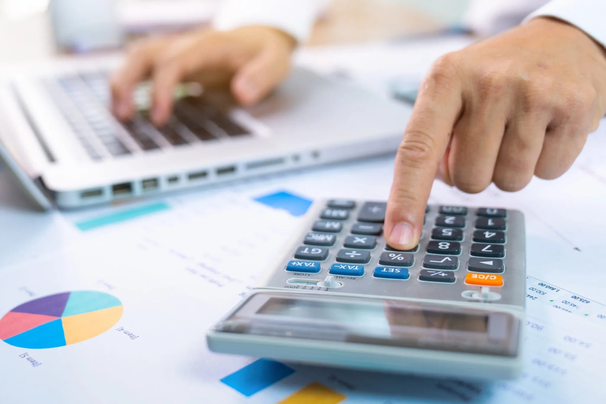 يد شخص يستخدم آلة حاسبة بجانب كمبيوتر محمول مع مستندات مالية وأوراق بيانية، تعبير عن المحاسبة وإدارة البيانات المالية للشركات الصغيرة.