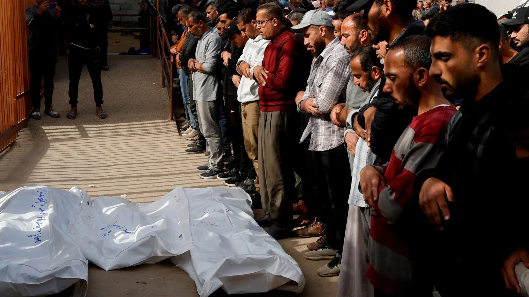 تجمع حشد من الرجال في جنازة، يحيطون بجثتين مغطاتين بأكياس بيضاء، تعبيرات الحزن واضحة على وجوههم.