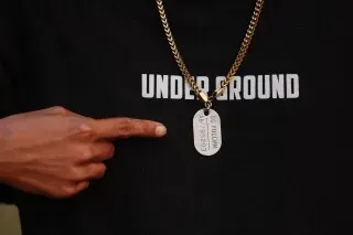 رجل يرتدي قميصًا مكتوبًا عليه "UNDER GROUND" ويشير بإصبعه إلى قلادة تحمل معلومات شخصية، تعكس تجارب العمل القاسية في الخارج.