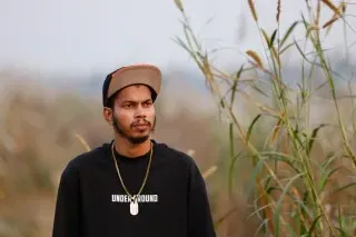 شاب بنغالي يقف في منطقة طبيعية، يرتدي سترة سوداء وقبعة، مع خلفية من الأعشاب الطويلة، يعبر عن مشاعر القلق والتفكير.