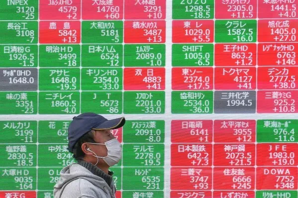 رجل يرتدي كمامة يسير بجانب لوحة تعرض أسعار الأسهم المتقلبة في اليابان، حيث تظهر الأرقام باللونين الأخضر والأحمر.