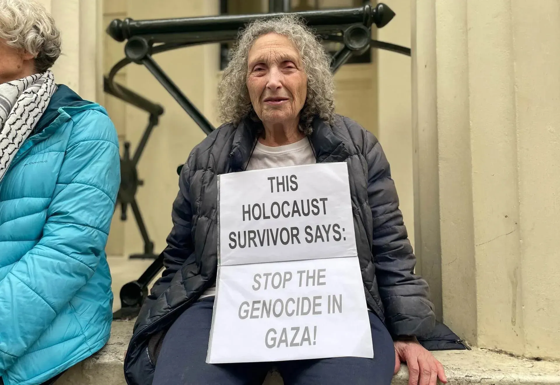 ناجية من الهولوكوست تحمل لافتة تطالب بوقف الإبادة الجماعية في غزة، تجلس في فعالية تذكارية بلندن.