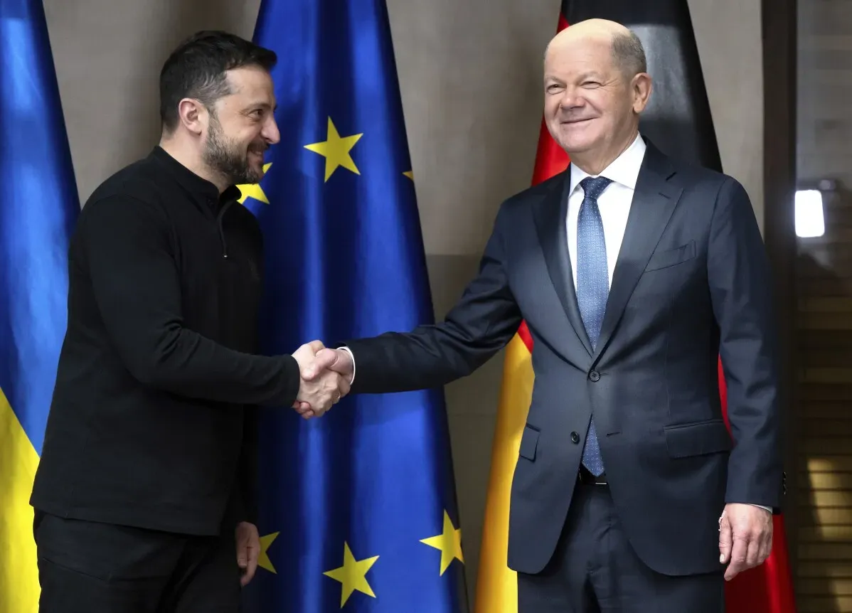 الرئيس الأوكراني فولوديمير زيلينسكي والمستشار الألماني أولاف شولتس يتصافحان في مؤتمر ميونيخ للأمن، مع أعلام أوكرانيا والاتحاد الأوروبي وخلفهما.